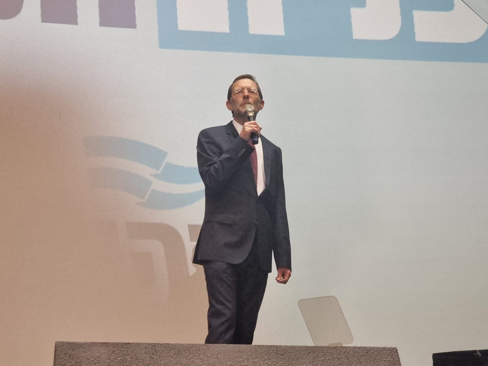 משה פייגלין נואם על הבמה עם מיקרופון, על רקע מיתוג זהות